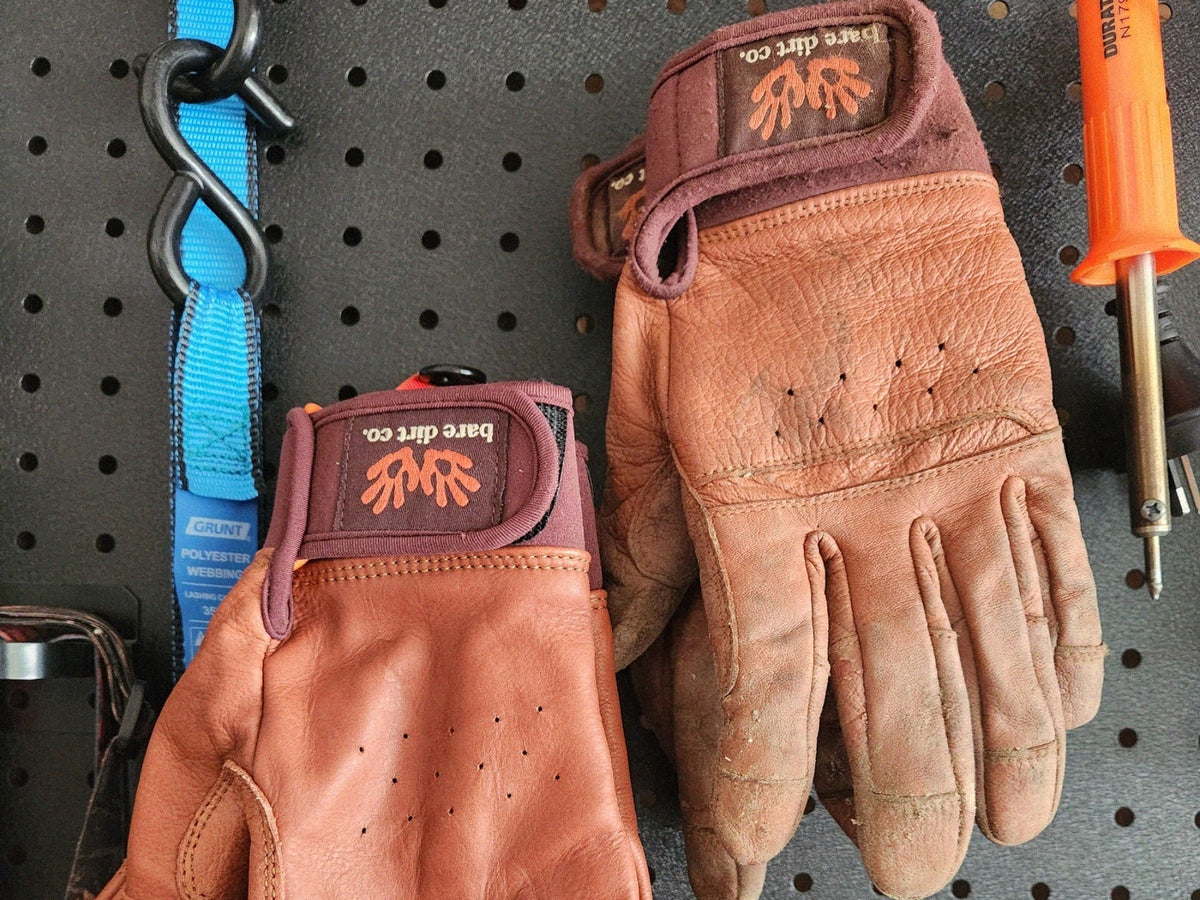 A pair of Bare Dirt Co. leather work gloves displayed on a dark gray pegboard, alongside a blue strap and an orange-handled tool.