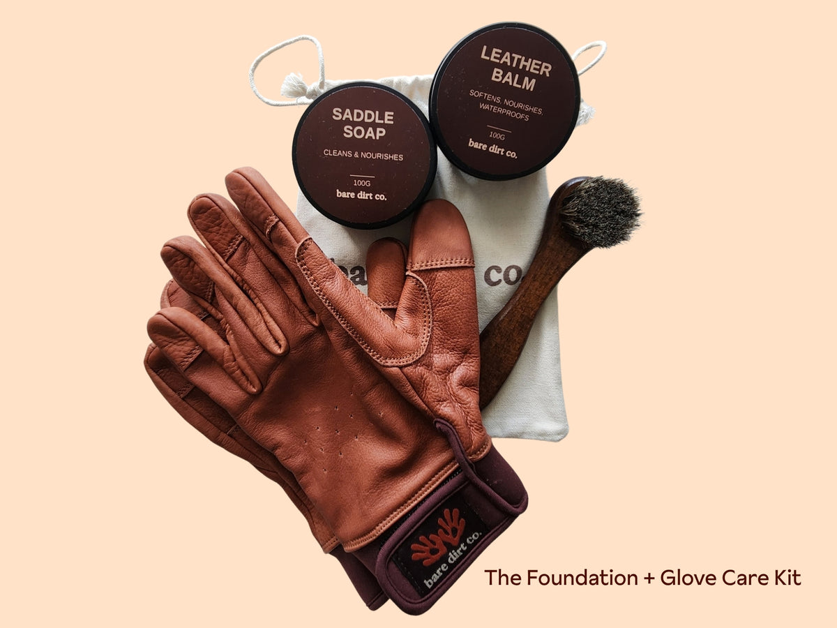 Glove care kit with a brown leather glove, two black lids labeled 'Saddle Soap' and 'Leather Balm', a white drawstring pouch, and a brush on a beige background. Text at bottom reads 'The Foundation + Glove Care Kit'.